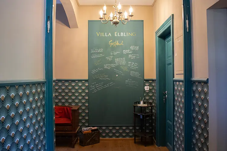 Ein gemütlicher Flur mit tealfarbener Vertäfelung und einer Kreidetafel mit der Aufschrift "Villa Elbling Gästebuch", die mit handgeschriebenen Gästebucheinträgen versehen ist. Eine kleine Holzbank und ein runder Tisch stehen unter einem goldenen Kronleuchter.