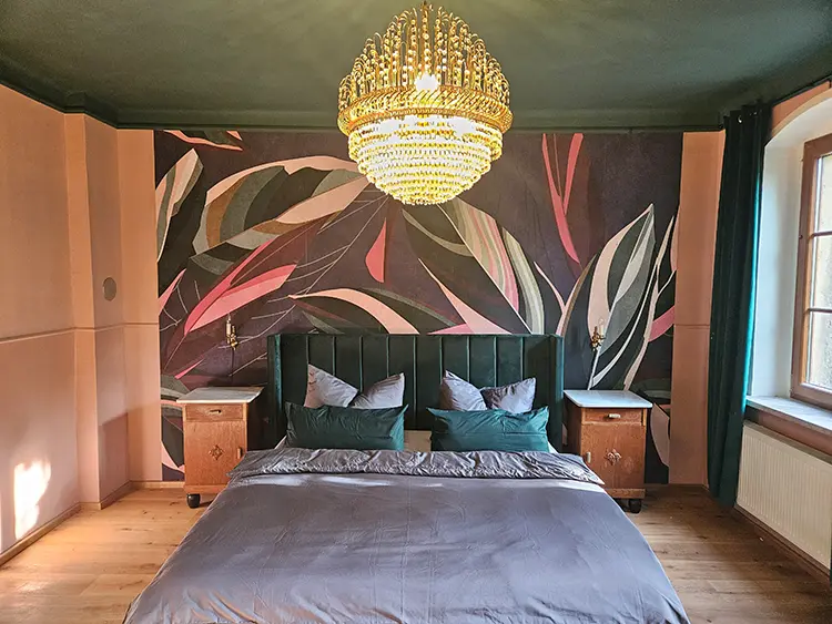 Ein stilvolles Schlafzimmer mit einem großen Bett, einem grünen Kopfteil, grauer Bettwäsche, zwei hölzernen Nachttischen, einer Akzentwand mit Blättermuster, einem funkelnden Kronleuchter und Sonnenlicht, das durch ein Fenster fällt.
