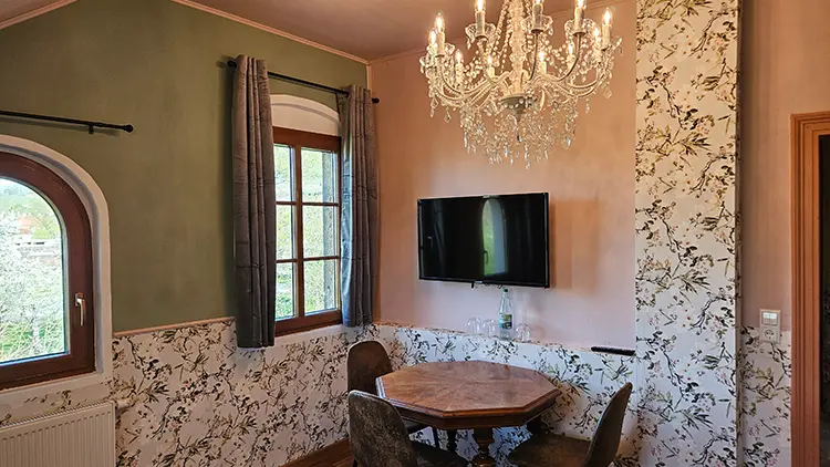 Ein kleiner Essbereich mit einem runden Tisch und vier Stühlen befindet sich unter einem Kristalllüster. Der Raum hat eine Blumentapete, grün und beige gestrichene Wände, zwei Fenster mit Vorhängen und einen Flachbildfernseher an der Wand.