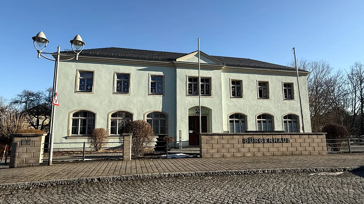 Ein zweistöckiges, hellgelbes Gebäude mit Bogenfenstern und einem Ziegeldach, das mit "Bürgerhaus" beschriftet ist, steht unter einem klaren blauen Himmel. Eine Straßenlaterne und spärliche Winterbäume sind rund um den kopfsteingepflasterten Bürgersteig zu sehen.