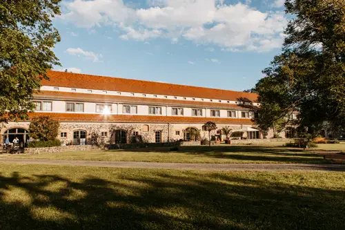 Ein großes Steingebäude mit rotem Ziegeldach befindet sich hinter einer Rasenfläche und Bäumen, die in warmes Sonnenlicht getaucht sind. Dieser historische Ort ist perfekt für eine Hochzeit am Gut am See und bietet mit seinen gewölbten Türen und einladenden Fenstern einen friedlichen Charme.