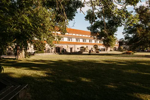 Ein großes Steingebäude mit rotem Ziegeldach befindet sich hinter einer Rasenfläche und Bäumen, die in warmes Sonnenlicht getaucht sind. Dieser historische Ort ist perfekt für eine Hochzeit am Gut am See und bietet mit seinen gewölbten Türen und einladenden Fenstern einen friedlichen Charme.