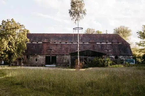 Eine rustikale, verwitterte Scheune mit rotem Ziegeldach steht auf einer grasbewachsenen Wiese, perfekt für eine Hochzeit auf Gut am See. Umgeben von grünen Bäumen und unter einem teilweise bewölkten Himmel verleihen ein hoher Baum und eine runde Metallkonstruktion der Szene Charme.