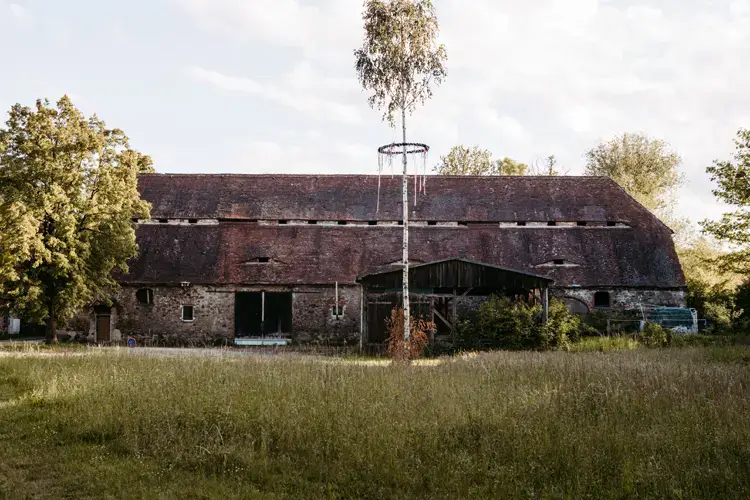 Eine rustikale, verwitterte Scheune mit rotem Ziegeldach steht auf einer grasbewachsenen Wiese, perfekt für eine Hochzeit auf Gut am See. Umgeben von grünen Bäumen und unter einem teilweise bewölkten Himmel verleihen ein hoher Baum und eine runde Metallkonstruktion der Szene Charme.