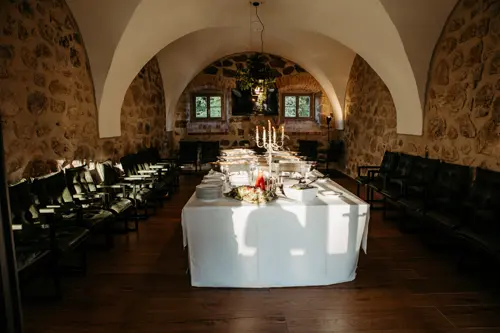 Ein Buffet in einem rustikalen Raum mit Steinwänden, gedeckt für eine Hochzeit am Gut am See, bietet Teller, Salate, Schüsseln mit Speisen und einen beleuchteten Kandelaber als Mittelstück unter gewölbten Decken mit natürlichem Licht aus zwei Fenstern.