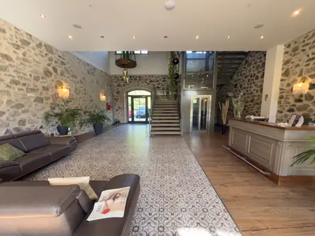 Eine geräumige Hotellobby mit Steinwänden, gemusterten Fliesen, braunen Ledersofas, einem hölzernen Empfangstresen, großen Topfpflanzen und einer zentralen Treppe - perfekt für Gäste, die eine Hochzeit am Gut am See besuchen.