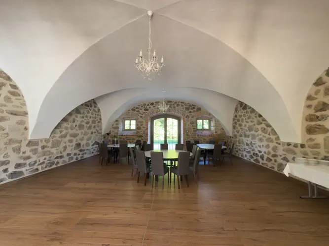 Ein geräumiger Raum mit Steinmauern, gewölbten Decken, einem Kronleuchter, Holzböden und mehreren runden Tischen - perfekt für eine Hochzeit am Gut am See - mit Stühlen, die in der Nähe von großen Fenstern angeordnet sind, die Tageslicht hereinlassen.