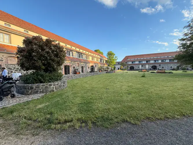 Ein weiter Blick auf ein großes Steingebäude mit rotem Dach in Gut am See, perfekt für eine Hochzeit am Gut am See, umgeben von einer großen grünen Wiese, Bäumen und einem klaren blauen Himmel. Sitzgelegenheiten im Freien sind in der Nähe des Gebäudes zu sehen.