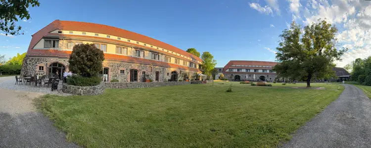 Ein weiter Blick auf ein großes Steingebäude mit rotem Dach in Gut am See, perfekt für eine Hochzeit am Gut am See, umgeben von einer großen grünen Wiese, Bäumen und einem klaren blauen Himmel. Sitzgelegenheiten im Freien sind in der Nähe des Gebäudes zu sehen.