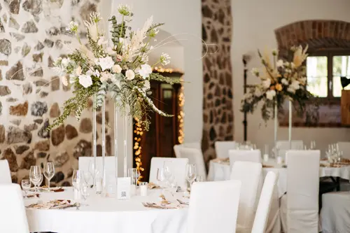 Eleganter Hochzeitsempfang mit runden Tischen, die mit weißen Tüchern bedeckt sind, hohen Mittelstücken aus weißen und grünen Blumen und ordentlich arrangierten Gläsern an einem rustikalen Ort mit Steinmauern - perfekt für eine Hochzeit am Gut am See.