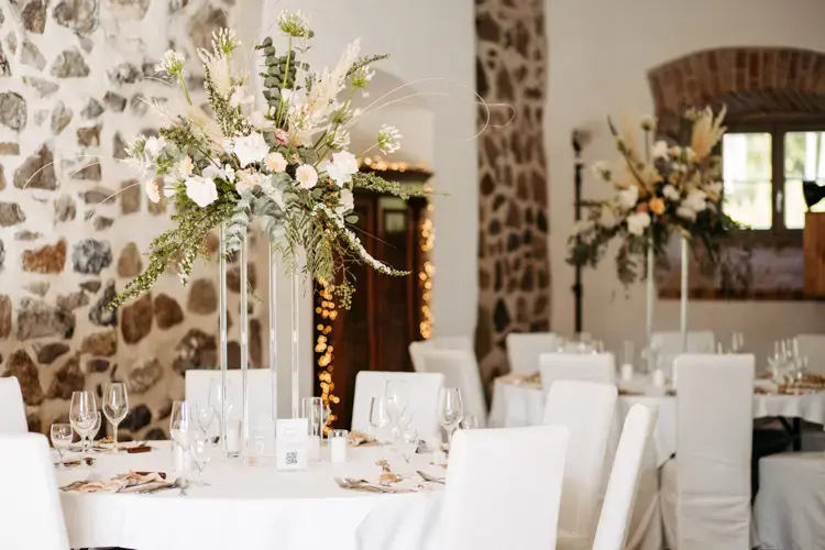 Eleganter Hochzeitsempfang mit runden Tischen, die mit weißen Tüchern bedeckt sind, hohen Mittelstücken aus weißen und grünen Blumen und ordentlich arrangierten Gläsern an einem rustikalen Ort mit Steinmauern - perfekt für eine Hochzeit am Gut am See.