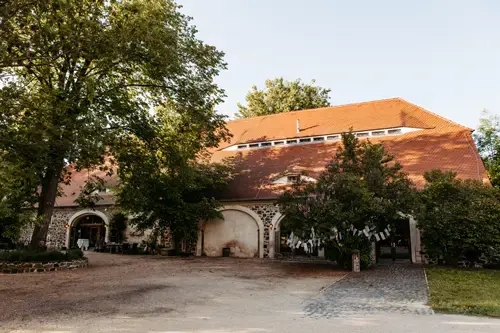 Ein großes, rustikales Gebäude mit rotem Ziegeldach und gewölbten Türen, das sich perfekt für eine Hochzeit am Gut am See eignet, wird an einem sonnigen Tag teilweise von hohen grünen Bäumen beschattet. Ein gepflasterter Weg und ein unbefestigter Boden führen zum Eingang.