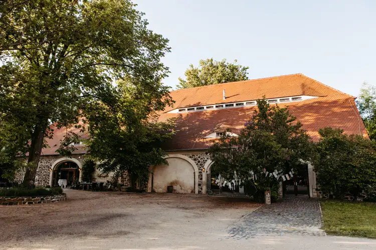 Ein großes, rustikales Gebäude mit rotem Ziegeldach und gewölbten Türen, das sich perfekt für eine Hochzeit am Gut am See eignet, wird an einem sonnigen Tag teilweise von hohen grünen Bäumen beschattet. Ein gepflasterter Weg und ein unbefestigter Boden führen zum Eingang.