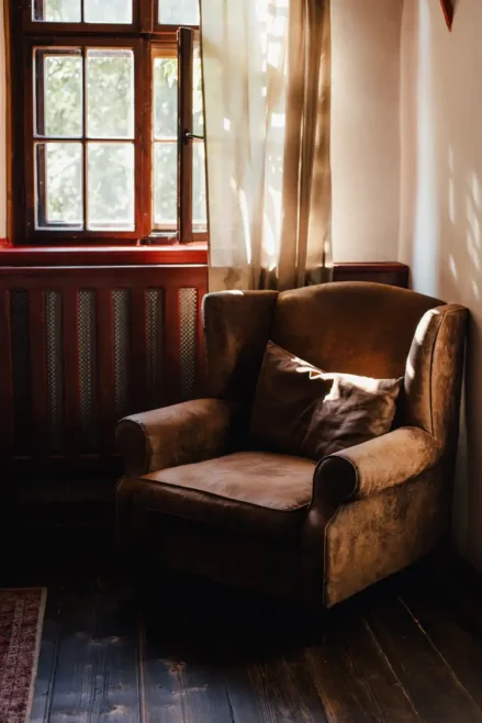 Ein gemütlicher brauner Sessel mit Kissen steht in einer sonnenbeschienenen Ecke an einem Fenster mit transparenten Vorhängen, die sanftes Licht und Schatten auf den dunklen Holzboden werfen.