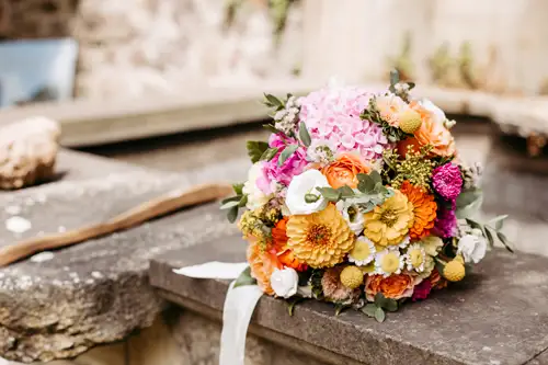 Ein farbenfroher Blumenstrauß mit rosa, orangefarbenen, gelben und weißen Blüten liegt auf einem Steinsockel im Freien, mit einem rustikalen Hintergrund, der leicht unscharf ist.