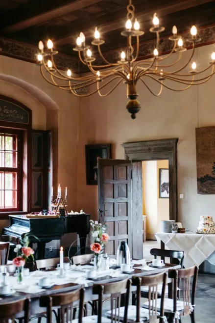 Ein warm beleuchteter Speisesaal mit einem langen Tisch, der für eine Mahlzeit gedeckt ist, mit Holzstühlen, einem Kronleuchter, frischen Blumen, Kerzen und einem Klavier an einem großen Fenster, durch das Sonnenlicht fällt.