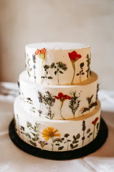 Eine dreistöckige weiße Torte, dekoriert mit gepressten bunten essbaren Blumen und Grünzeug, auf einer schwarzen Tortenplatte.