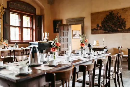 Ein langer hölzerner Esstisch mit Tassen, Untertassen, Wasserkochern und Blumenvasen steht in einem warm beleuchteten Raum im Vintage-Stil mit Holzstühlen, großen Fenstern und klassischem Dekor.