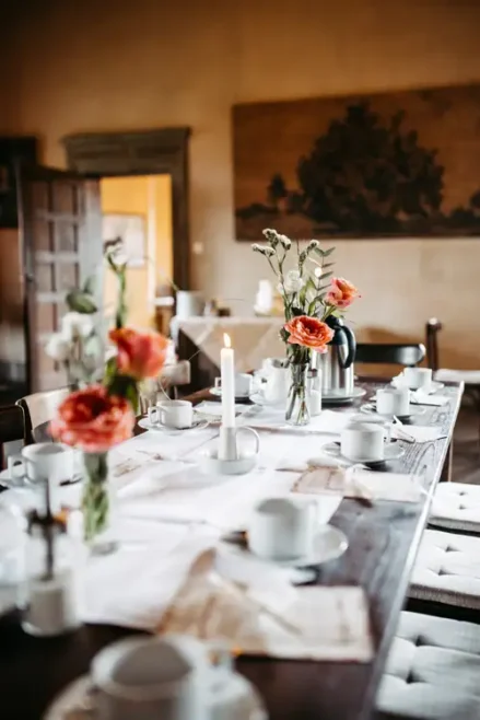 Ein hölzerner Esstisch, gedeckt für eine Mahlzeit, dekoriert mit weißem Geschirr, Tassen, Silberbesteck, brennenden Kerzen, orangefarbenen und weißen Blumen in Glasvasen, mit natürlichem Licht, das durch eine offene Tür im Hintergrund einfällt.