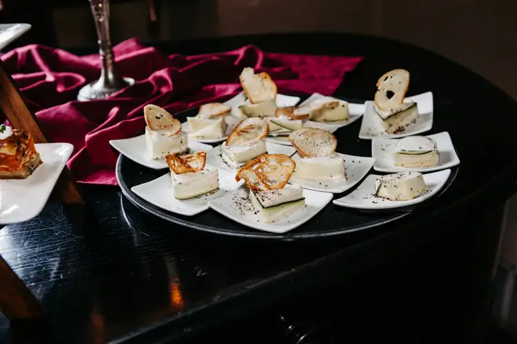 Kleine quadratische Teller mit Gourmet-Häppchen, jeweils mit einer Scheibe Käse und einer dünnen Scheibe Brot oder Cracker, die auf einem dunklen runden Tisch mit einem weinroten Tuch im Hintergrund angeordnet sind.