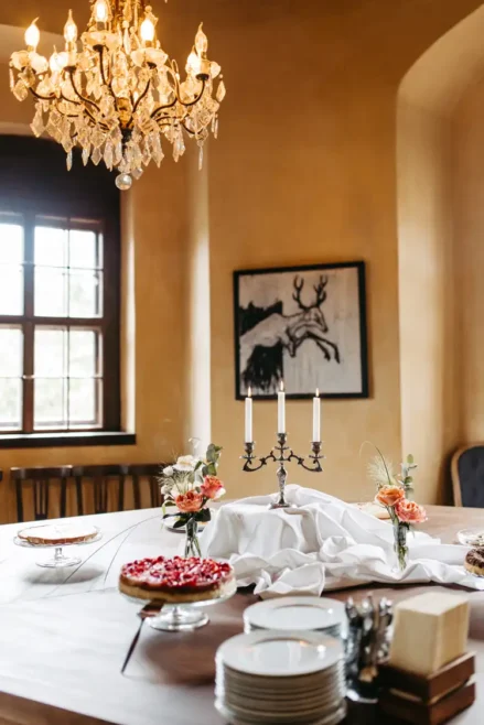 Ein eleganter Esstisch mit Desserts, Blumen und Tellern unter einem beleuchteten Kristalllüster in einem warm beleuchteten Raum mit einem Fenster und gerahmter Kunst an der Wand.