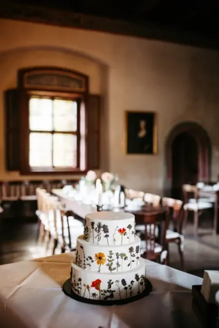Eine dreistöckige weiße Torte, verziert mit bunten gepressten Blumen, steht auf einem Tisch in einem eleganten, sanft beleuchteten Raum mit Holzstühlen, Tischen und einem großen Fenster im Hintergrund.