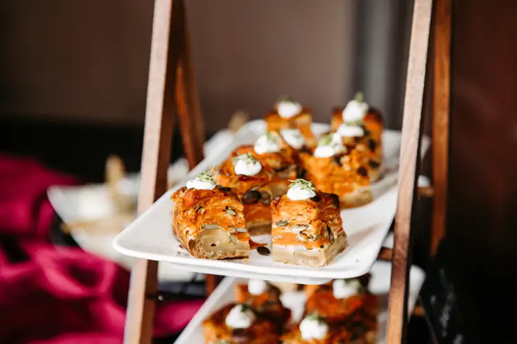 Nahaufnahme einer weißen, rechteckigen Platte mit mehreren Stücken herzhaften Gebäcks, belegt mit Kräutern und einem Klecks Sahne, die auf einem gestuften Holzständer präsentiert werden. Der Hintergrund ist sanft verschwommen.