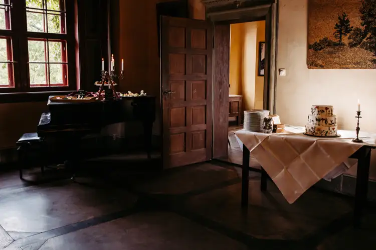 Ein warm beleuchteter Raum mit einem für eine Feier gedeckten Tisch, auf dem ein gestapelter Kuchen, gestapelte Teller und eine einzelne brennende Kerze stehen. Eine offene Holztür gibt den Blick auf einen weiteren, sanft beleuchteten Raum im Hintergrund frei.