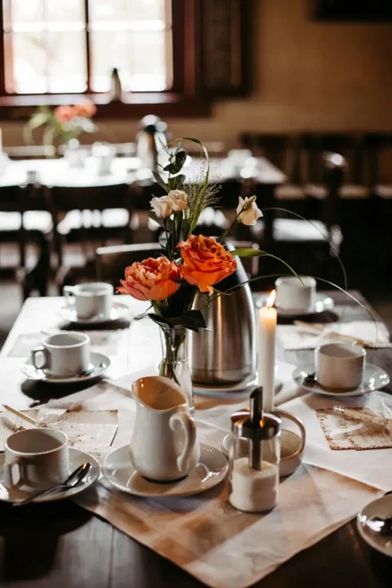 Ein gemütlich gedeckter Teetisch mit weißen Tassen, Untertassen, einer Teekanne, einer Kerze und einer Vase mit frischen Blumen, in einem warm beleuchteten Raum mit Holzstühlen und einem Fenster im Hintergrund.