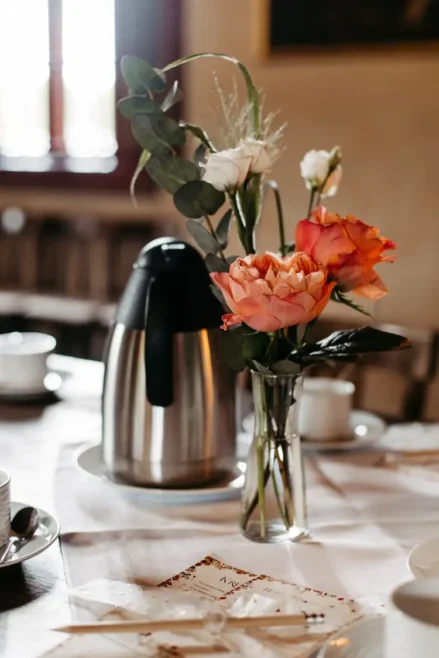 Eine Kaffeekaraffe aus Edelstahl steht auf einem Tisch neben einer Glasvase mit rosa und orangefarbenen Blumen, umgeben von Teetassen und Untertassen in einem sanft beleuchteten Raum.