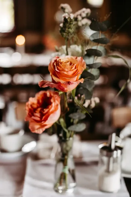Eine Glasvase mit orangefarbenen Rosen und Grünzeug steht auf einem für eine Mahlzeit gedeckten Tisch, mit Tassen, Untertassen und einem Zuckerspender, die im Hintergrund verschwimmen.