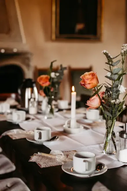 Ein mit weißen Tassen und Untertassen gedeckter Esstisch, eine brennende Kerze, rosa Rosen in Glasvasen und eine Kaffeekanne schaffen eine elegante, gemütliche Atmosphäre.