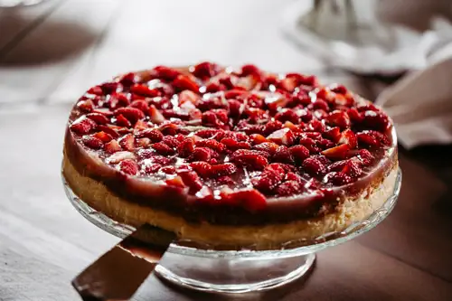 Ein Käsekuchen mit einer Schicht glasierter frischer Erdbeeren liegt auf einem gläsernen Kuchenständer, unter einer Scheibe befindet sich ein Servierspatel.