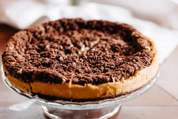 Ein runder Schokoladenstreuselkuchen mit einer dicken Kruste steht auf einem gläsernen Kuchenständer, teilweise aufgeschnitten, mit einem verschwommenen weißen Tuch im Hintergrund.