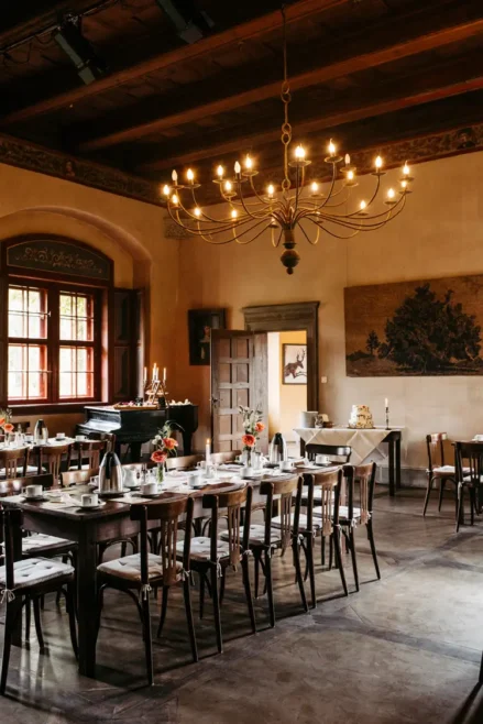 Ein warm beleuchteter Speisesaal mit Holztischen und -stühlen, die für eine Mahlzeit gedeckt sind. Blumenschmuck, ein Flügel, ein großer Kronleuchter und Sonnenlicht, das durch hohe Fenster einfällt, schaffen eine einladende Atmosphäre.