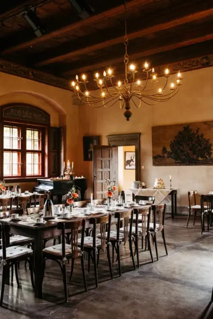 Ein warm beleuchteter Speisesaal mit Holztischen und -stühlen, die für eine Mahlzeit gedeckt sind. Blumenschmuck, ein Flügel, ein großer Kronleuchter und Sonnenlicht, das durch hohe Fenster einfällt, schaffen eine einladende Atmosphäre.