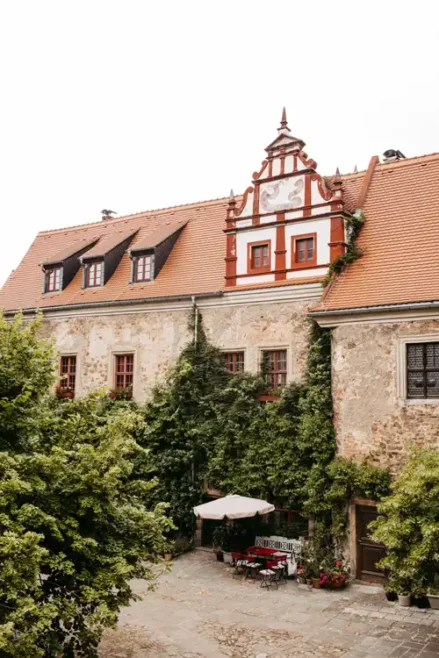 Historisches Steingebäude mit rotem Ziegeldach, verschnörkeltem Giebel und Efeu an den Wänden. Im Innenhof darunter steht ein Tisch mit einem weißen Sonnenschirm, umgeben von Grün und Bäumen.