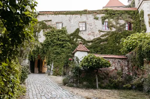Ein kopfsteingepflasterter Weg führt zu einem alten, mit grünem Efeu bewachsenen Steingebäude mit rot gerahmten Fenstern und einem kleinen bogenförmigen Eingang. Eine niedrige Steinmauer mit einem Ziegeldach und üppiges Grün umgeben den Eingang.