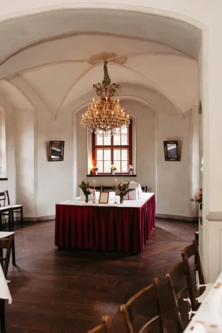 Ein formeller Speisesaal mit gewölbten Decken, einem großen Kronleuchter, einem für eine Veranstaltung gedeckten Tisch mit roten Röcken in der Mitte, Holzböden und einem von weißen Wänden eingerahmten Fenster.