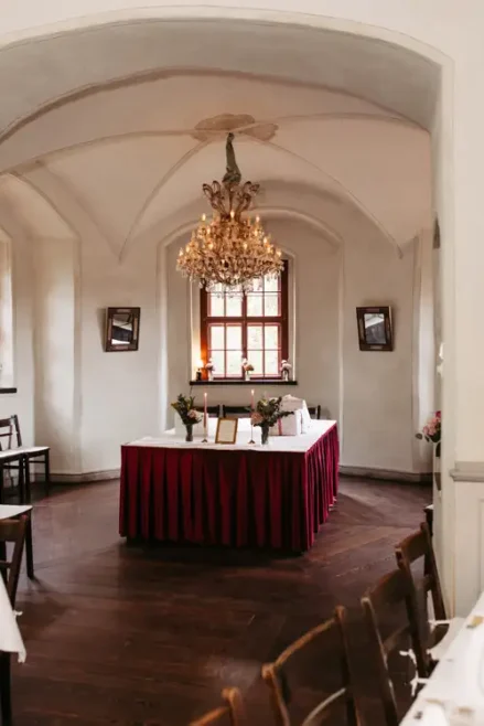 Ein formeller Speisesaal mit gewölbten Decken, einem großen Kronleuchter, einem für eine Veranstaltung gedeckten Tisch mit roten Röcken in der Mitte, Holzböden und einem von weißen Wänden eingerahmten Fenster.