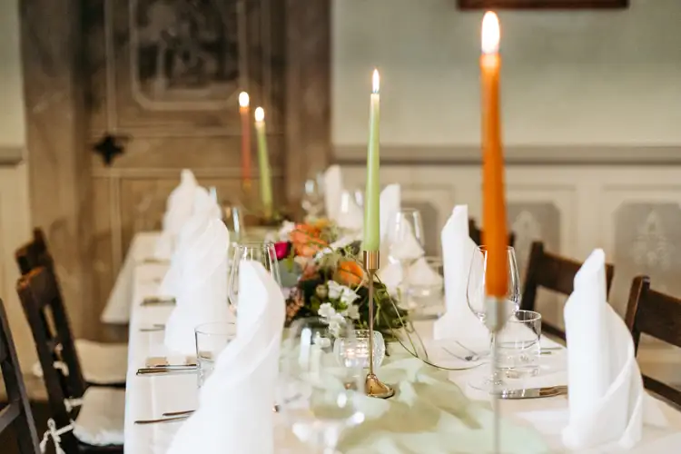 Ein formeller Esstisch ist mit weißen Servietten, Gläsern, Tellern und einem floralen Mittelstück gedeckt. Hohe, brennende Kerzen in Orange und Grün schaffen eine warme, elegante Atmosphäre. Auf beiden Seiten des Tisches stehen Holzstühle.