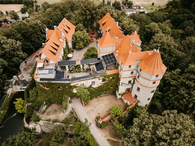 Luftaufnahme eines historischen Schlosses mit efeubewachsenen Steinmauern und orangefarbenen Ziegeldächern unter einem bewölkten Himmel, umgeben von Grün und Bäumen.