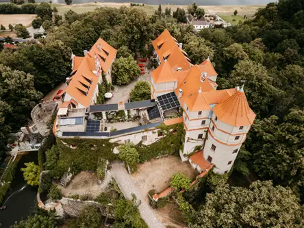 Luftaufnahme eines historischen Schlosses mit rot-orangefarbenen Dächern, umgeben von dichten grünen Bäumen und Landschaften, mit mehreren Türmen und einem Innenhof.