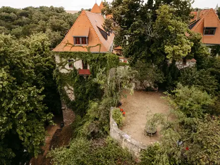 Luftaufnahme eines historischen Steingebäudes mit steilen orangefarbenen Dächern, teilweise mit grünem Efeu bewachsen, umgeben von dichten Bäumen und einem kleinen, ummauerten Hof.