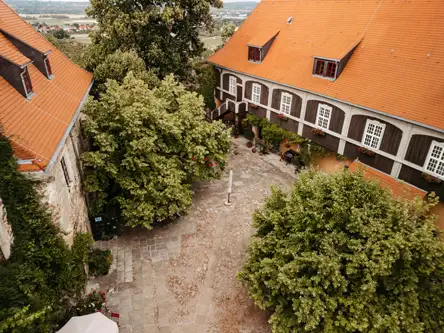 Luftaufnahme eines steinernen Innenhofs, umgeben von historischen Gebäuden mit orangefarbenen Ziegeldächern und weiß gestrichenen Fenstern, beschattet von großen grünen Bäumen.