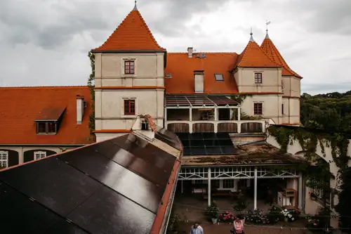 Ein großes, historisches Gebäude mit orangefarbenen Ziegeldächern, weißen Wänden und mehreren Türmen, von oben an einem bewölkten Tag betrachtet. Auf einem niedrigeren Dach im Vordergrund sind Sonnenkollektoren zu sehen.