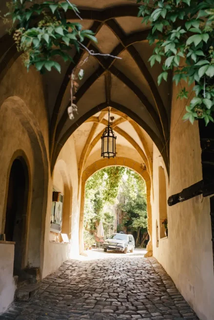 Ein steinerner Torbogen mit Gewölbe im gotischen Stil und einer hängenden Laterne öffnet sich zu einem sonnenbeschienenen Hof, in dem ein Auto geparkt ist.