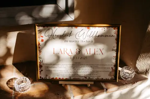 Ein gerahmtes Hochzeitsbegrüßungsschild in deutscher Sprache mit floraler Umrandung steht auf einem Sims zwischen zwei gläsernen Kerzenhaltern, die vom Sonnenlicht warm beleuchtet werden. Die Namen Lara & Alex und das Datum 11. Juli 2023 sind gut sichtbar angebracht.