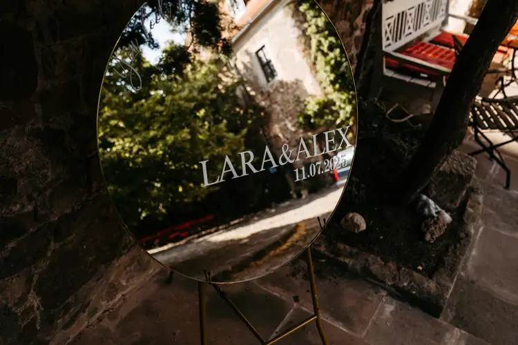 Ein rundes Spiegelschild auf einem Ständer reflektiert eine Szene im Freien und zeigt die Namen Lara & Alex mit dem Datum 11.07.2021 in weißer Schrift, was auf eine Hochzeitsfeier hindeutet.
