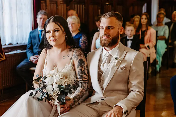 Braut und Bräutigam sitzen während einer Hochzeitszeremonie zusammen. Die Braut mit langen dunklen Haaren und Tattoos hält einen Strauß weißer Blumen in der Hand. Der Bräutigam, bärtig und in einem beigen Anzug aus dem Fotoshooting „Heiraten im Standesamt Tharandt“ von Melanie Kunert Fotografie, sitzt neben ihr, während die Gäste zuschauen.